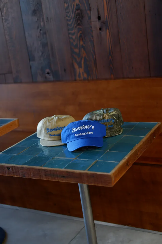 Blue, tan, and camouflage Rooster's caps on a blue tiled table