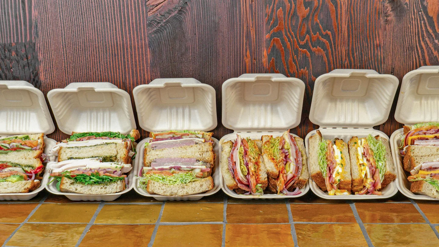 A lineup of Rooster's freshly made sandwiches in open takeout boxes along the tiled counter